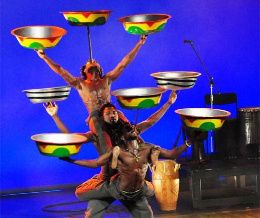 Drei Künstler balancieren Teller auf Stöcken und sitzen aufeinander auf dem Rücken